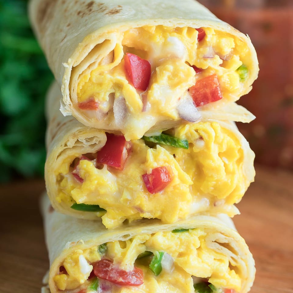 Three stacked breakfast burritos, filled with scrambled eggs, red peppers, and cheese, on a wooden board.