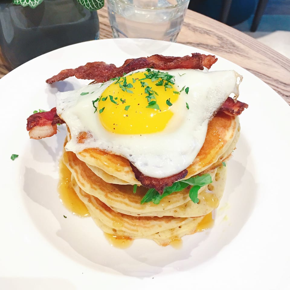 Stack of pancakes with bacon, a fried egg, and greens, drizzled with syrup on a white plate.