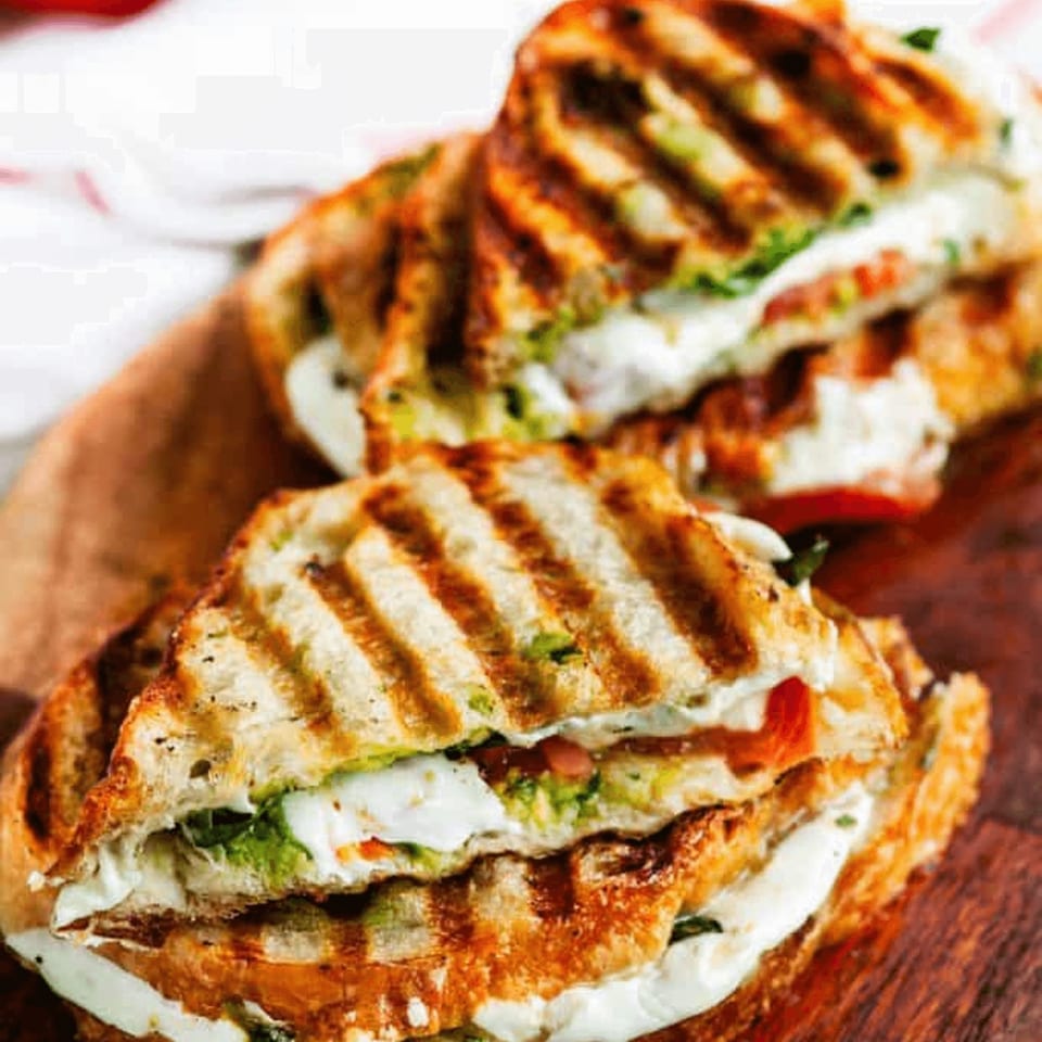 Two grilled paninis with melted cheese, tomatoes, and greens on a wooden board.