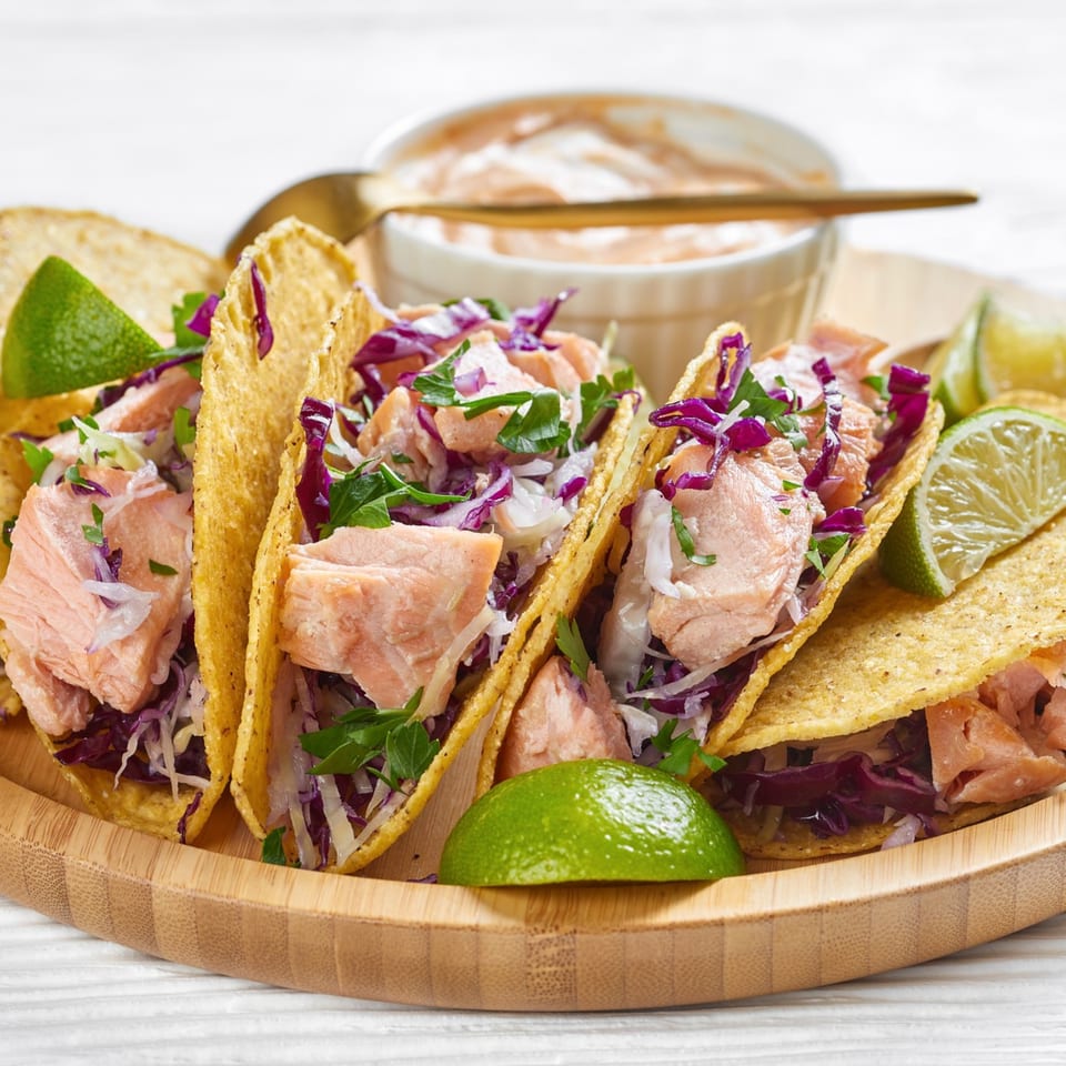 Four salmon tacos with red cabbage, cilantro, and lime wedges on a wooden plate.